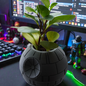 A round, panel-lined 3D printed planter holding a 4-inch leafy plant on a gaming desk with RGB lighting and figurines in the background
