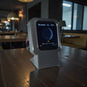 A compact 3D-printed moon phase display showing the current moon, countdown, weather, and time, sitting on a wooden table in a dimly lit room.
