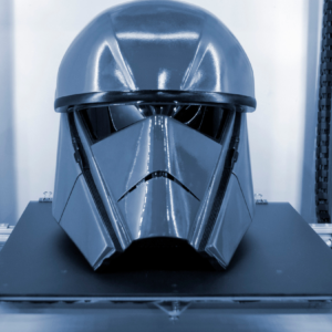 A large, glossy, angular sci-fi–style helmet sitting on the print bed of a professional 3D printer, surrounded by metal rails and cables.
